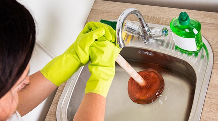 Gloved hands using a plunger in a kitchen sink